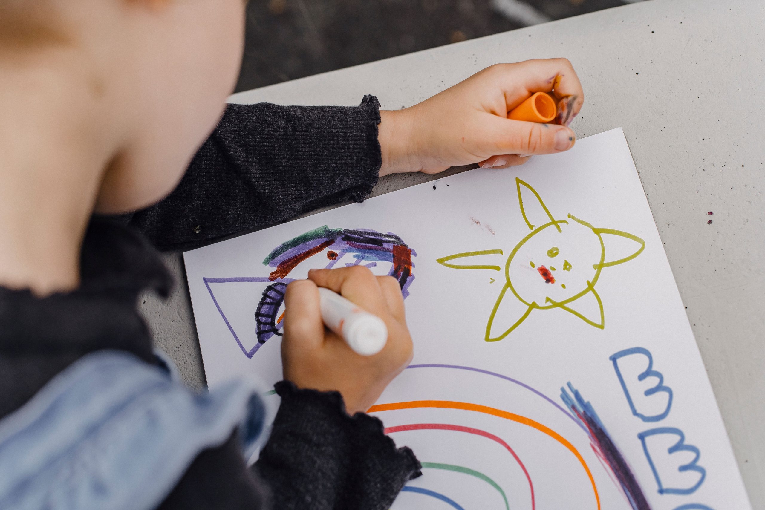 High angle of crop unrecognizable kid sitting at table and holding marker while painting colorful picture in album for drawing